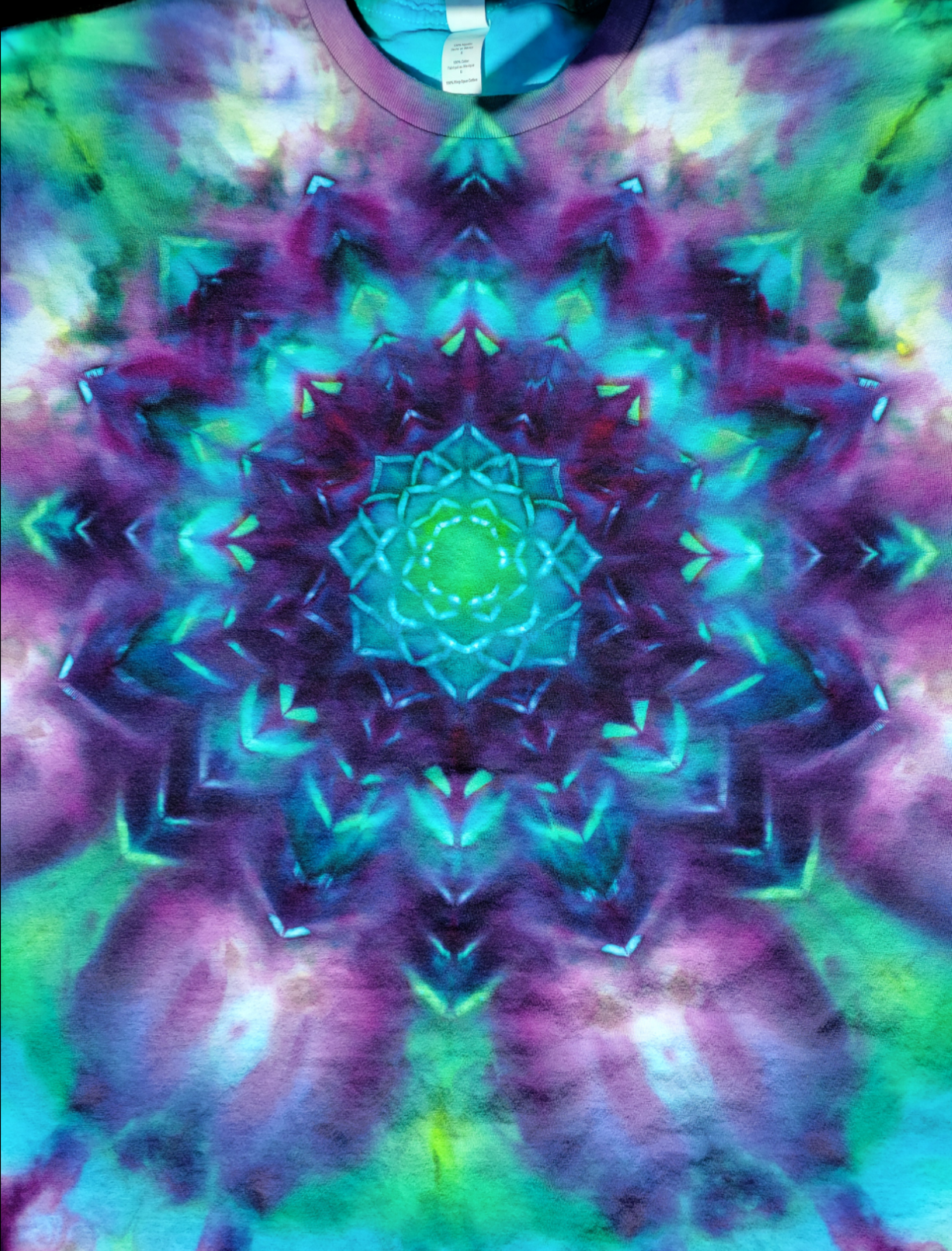 Close-up of Blooming Mandala pattern on front of tie-dyed t-shirt in purples, blues, and greens. Pattern resembles lotus blossom.